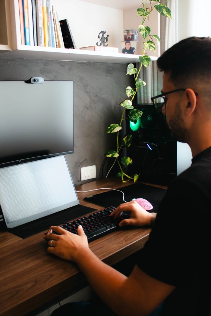 A young man working at a home office desk using a laptop and monitor, surrounded by tech and decor.